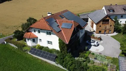 Aerial view of a house with solar panels on the roof, a garden and driveway, next to buildings, set against open fields.