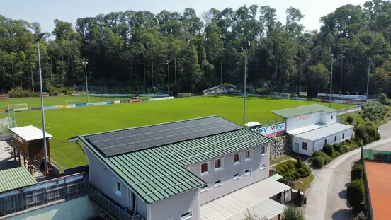 Sports field with soccer pitch, stands, and buildings with solar panels on the roof, surrounded by forest and paths