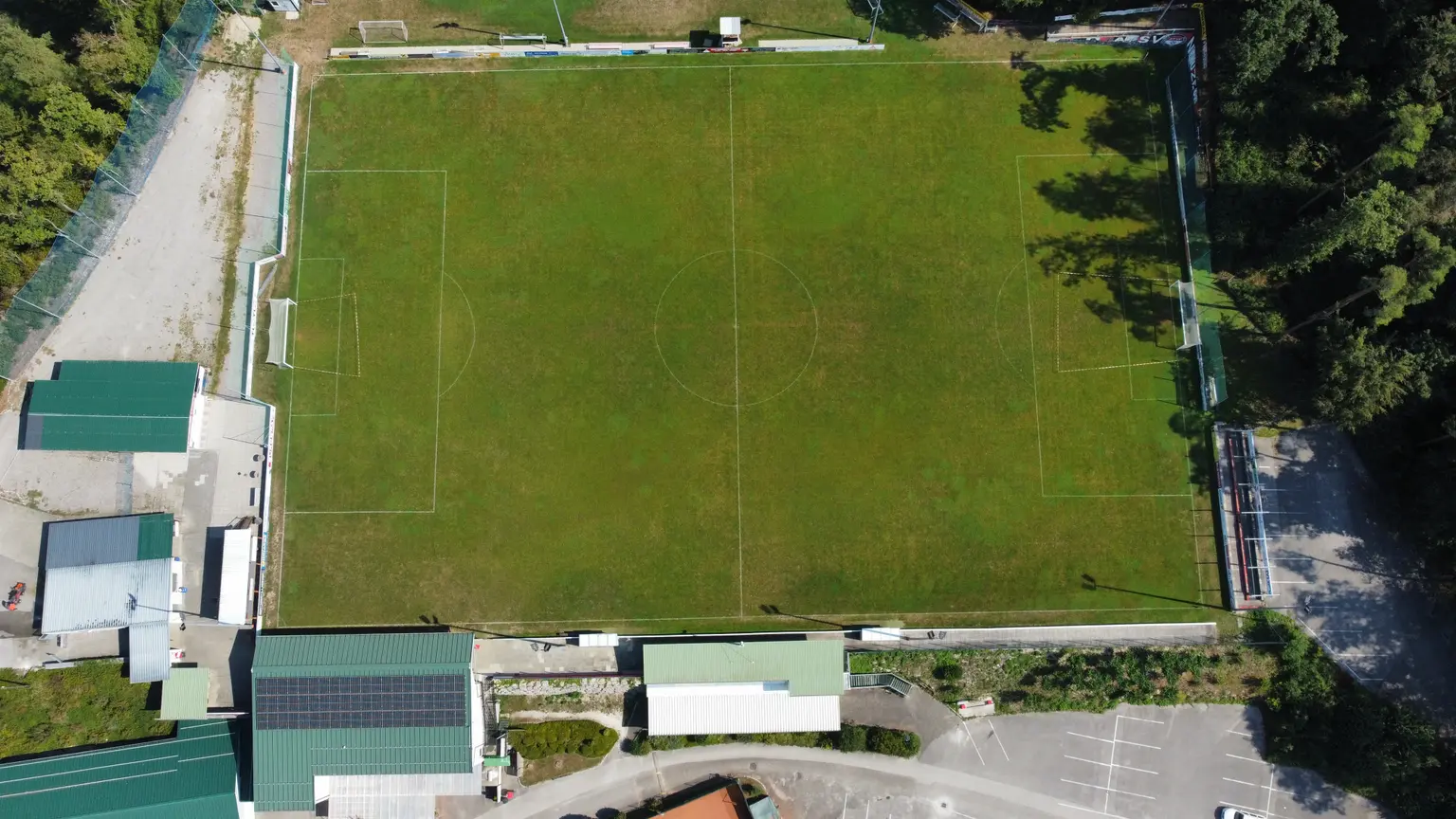 Football field from bird’s-eye view surrounded by forest, parking areas, and buildings with PV panels on the rooftops.