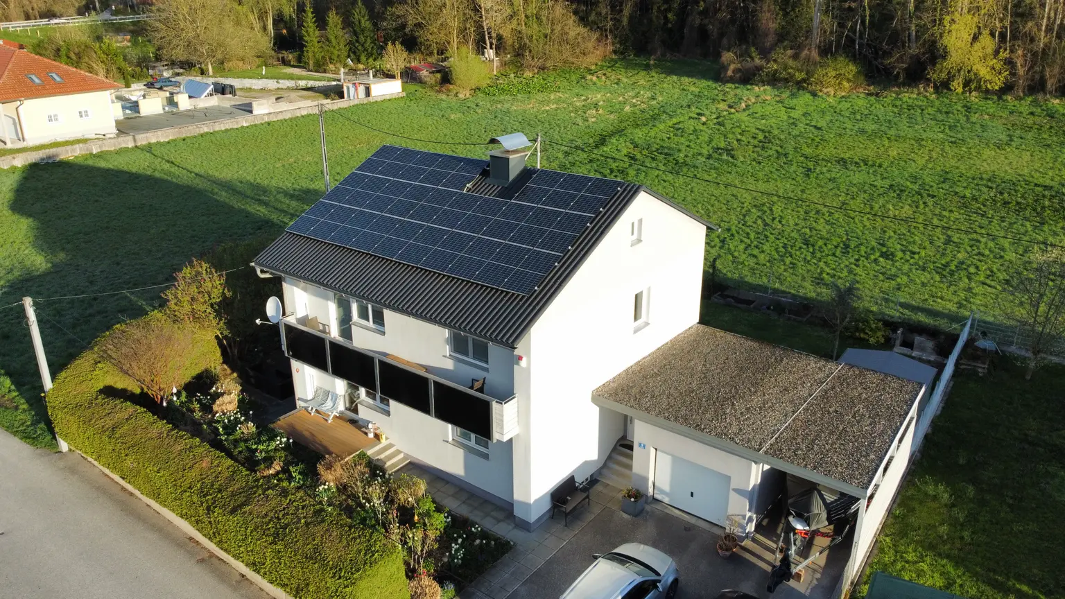House in late summer evening light with solar panels, a chimney on the roof, balcony, and garden fenced by shrubs.