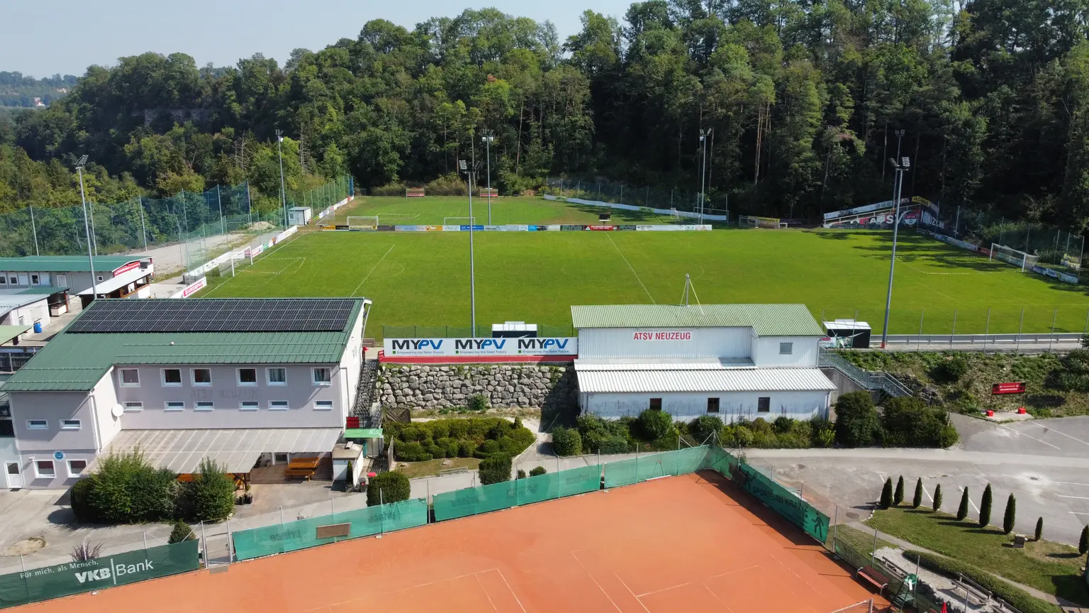 Aerial view of sports facility with soccer field, my-PV signage, tennis court, and buildings with rooftop solar panels.