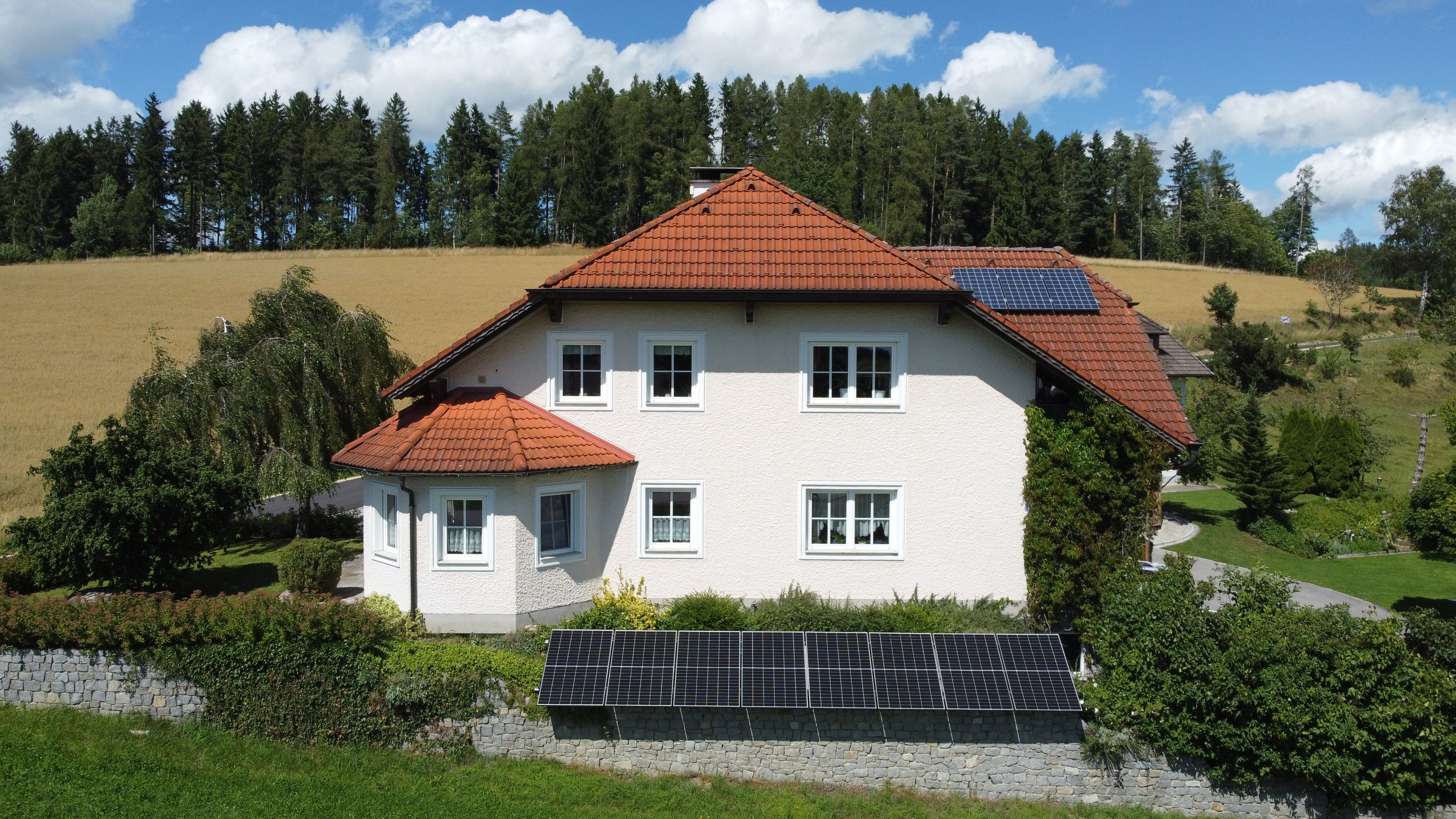 A white house with red roof tiles, solar panels, surrounded by greenery and trees, under a blue sky with clouds.
