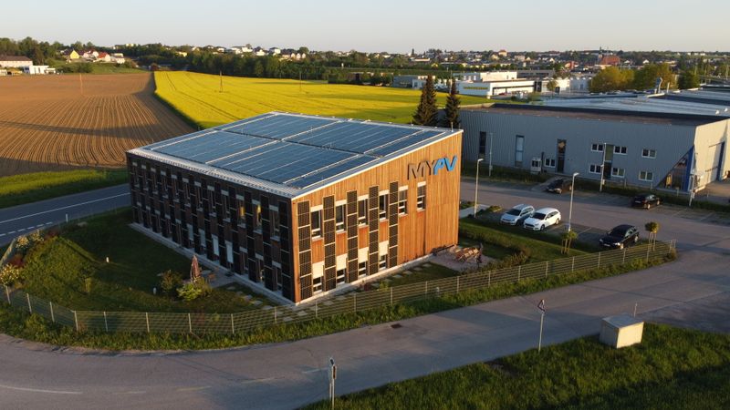 Aerial view of my-PV building with solar panels on roof and facade, visible logo, and cars in the parking lot.