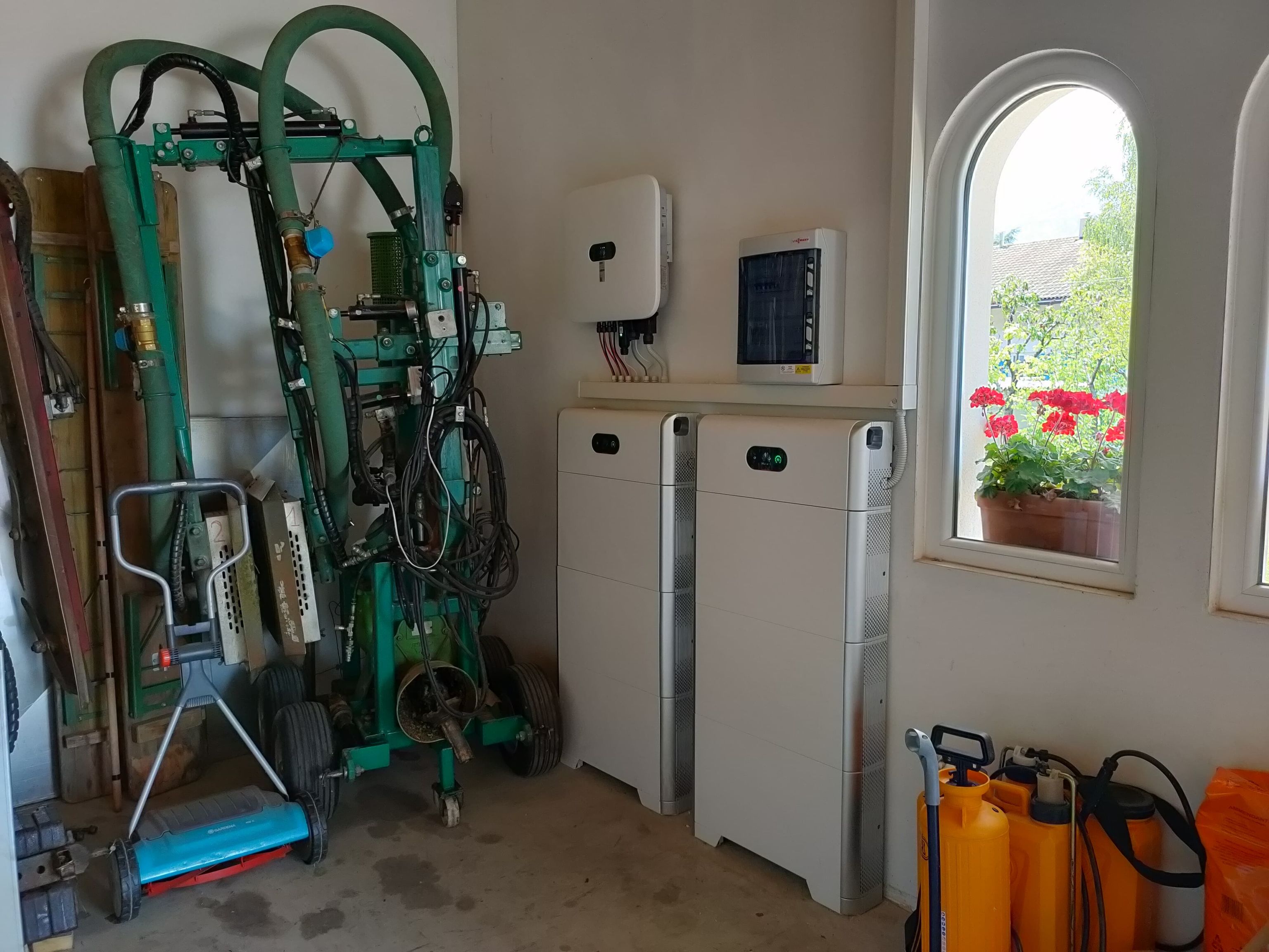 Technical room with two white batteries, control unit above, green equipment left, spray bottles and window with red flowers right.