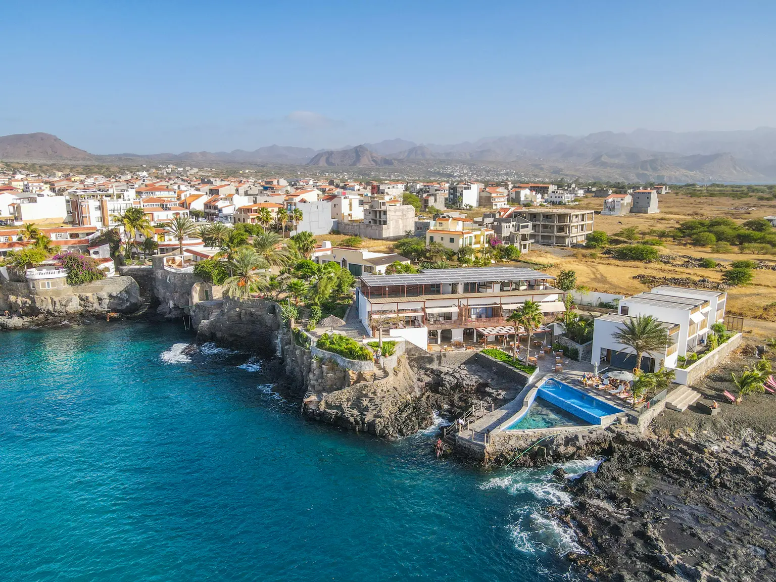 View from the sea of the hotel complex with pool by the water, other houses, palms, and shrubs in the background