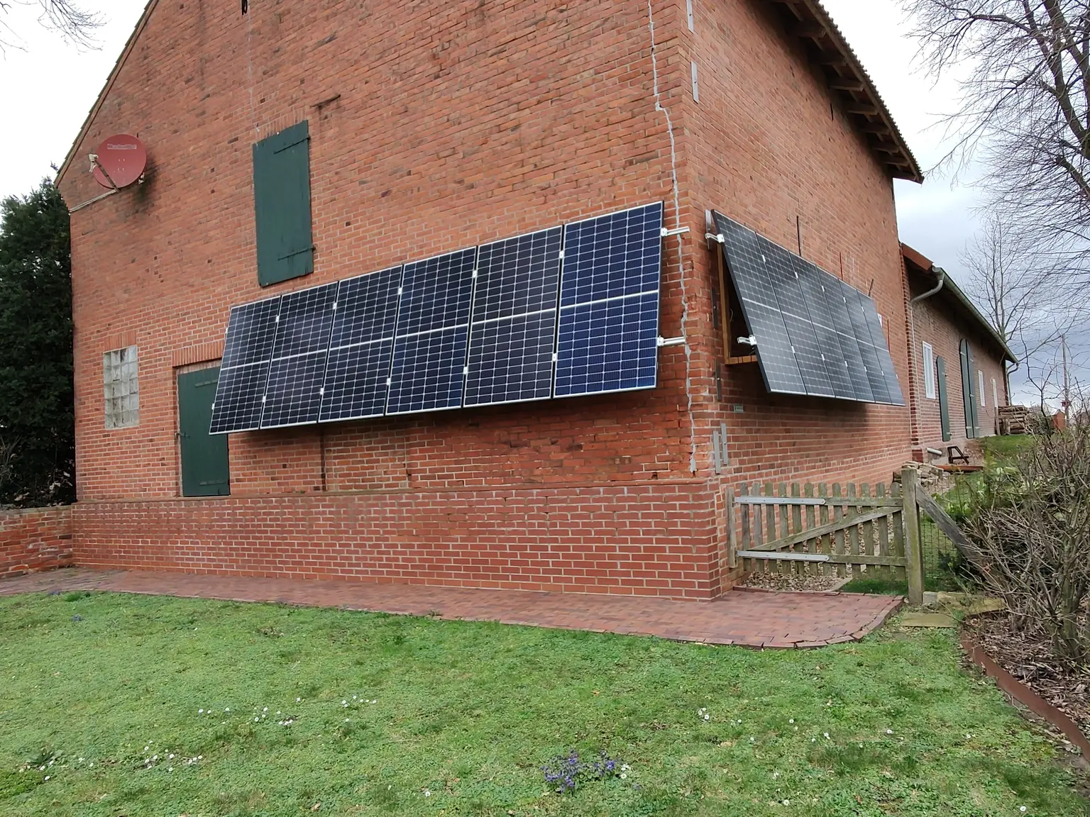 House with red brick wall and side-mounted PV panels, next to a garden with lawn and a fence along the side.