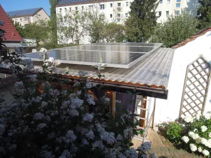 PV panels with 1.4 kWp on a small garden shed roof, with a large flowering plant in front.