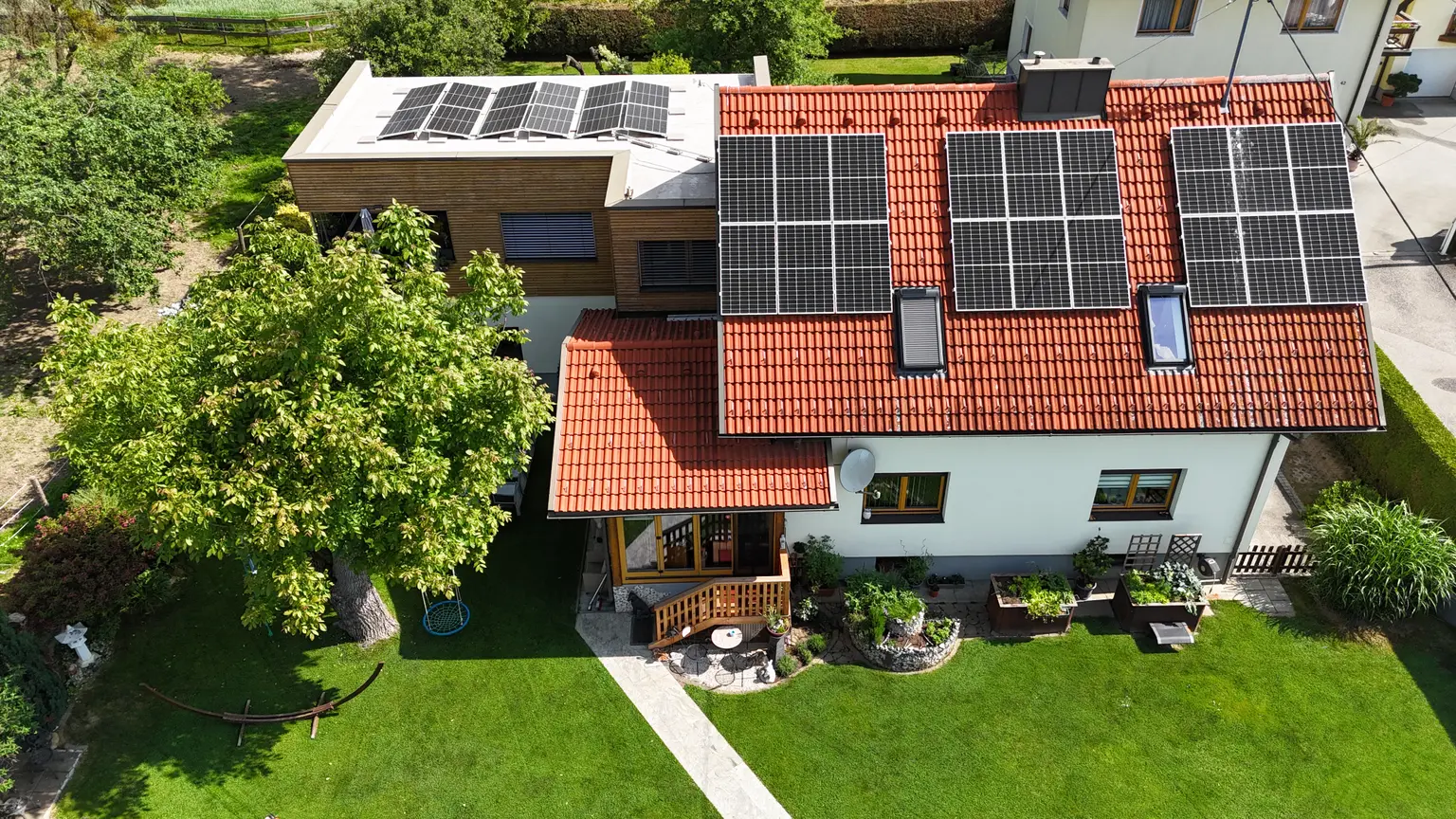 Aerial view of house with solar panels on red-tiled roof, surrounded by lush green garden and trees.