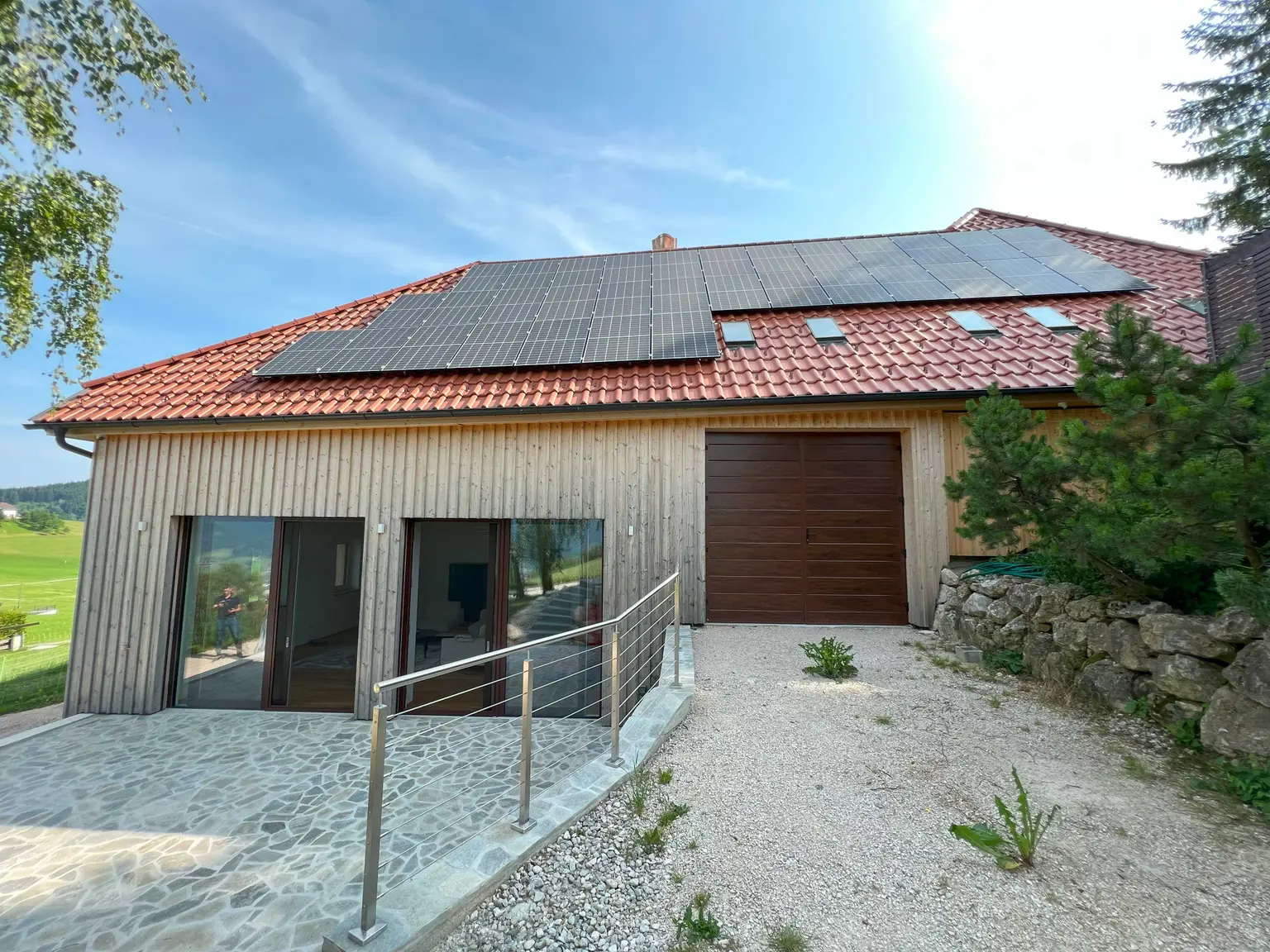 Farmhouse with wooden facade and PV panels on the roof, open glass balcony doors, and driveway leading to a garage.