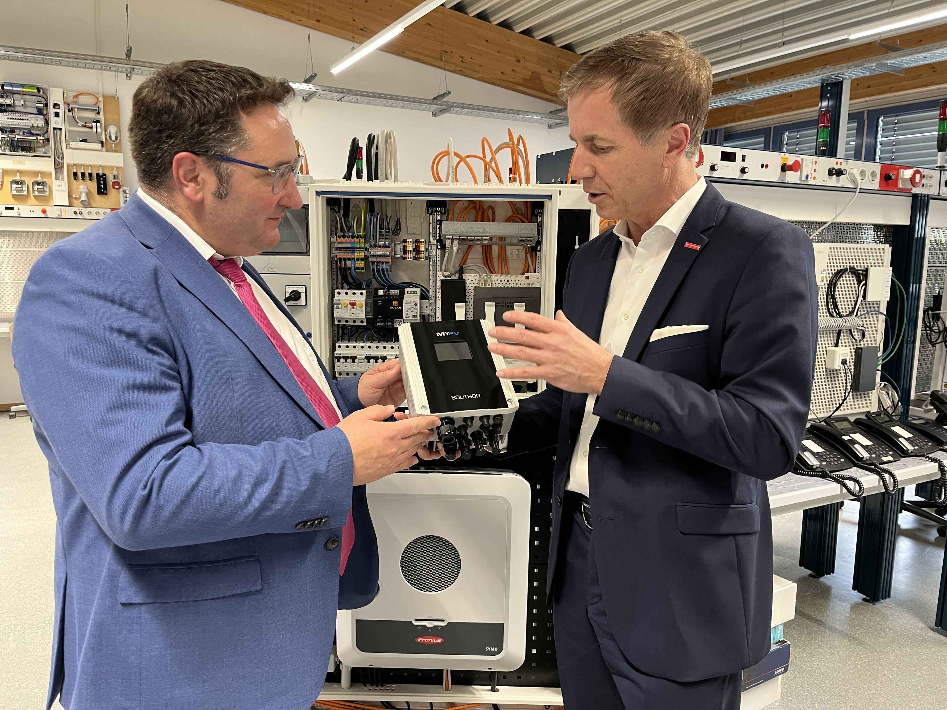 Two men in suits discuss an electronic device in a tech showroom, surrounded by wires and equipment displays.