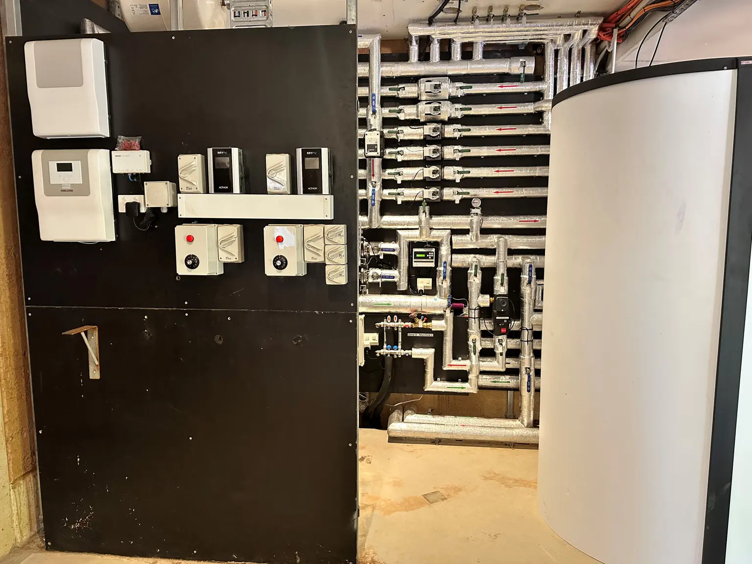 Room with control panel on left, complex wall pipes, and large cylindrical tank on right.