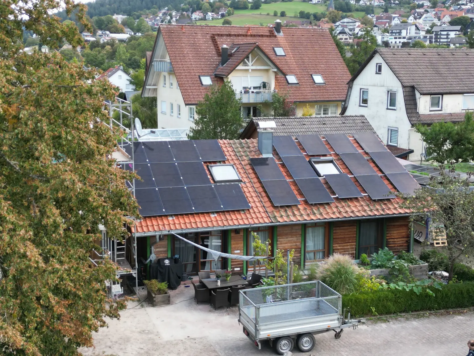 House with solar panels, wooden facade with green-framed windows, tree in front, and side structure for additions.