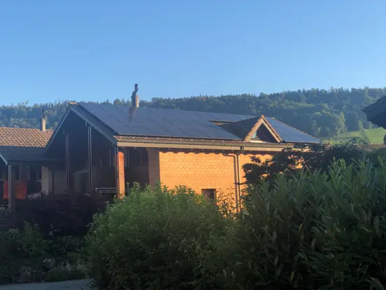 House with solar panels on the roof, surrounded by greenery and hills under a clear blue sky.
