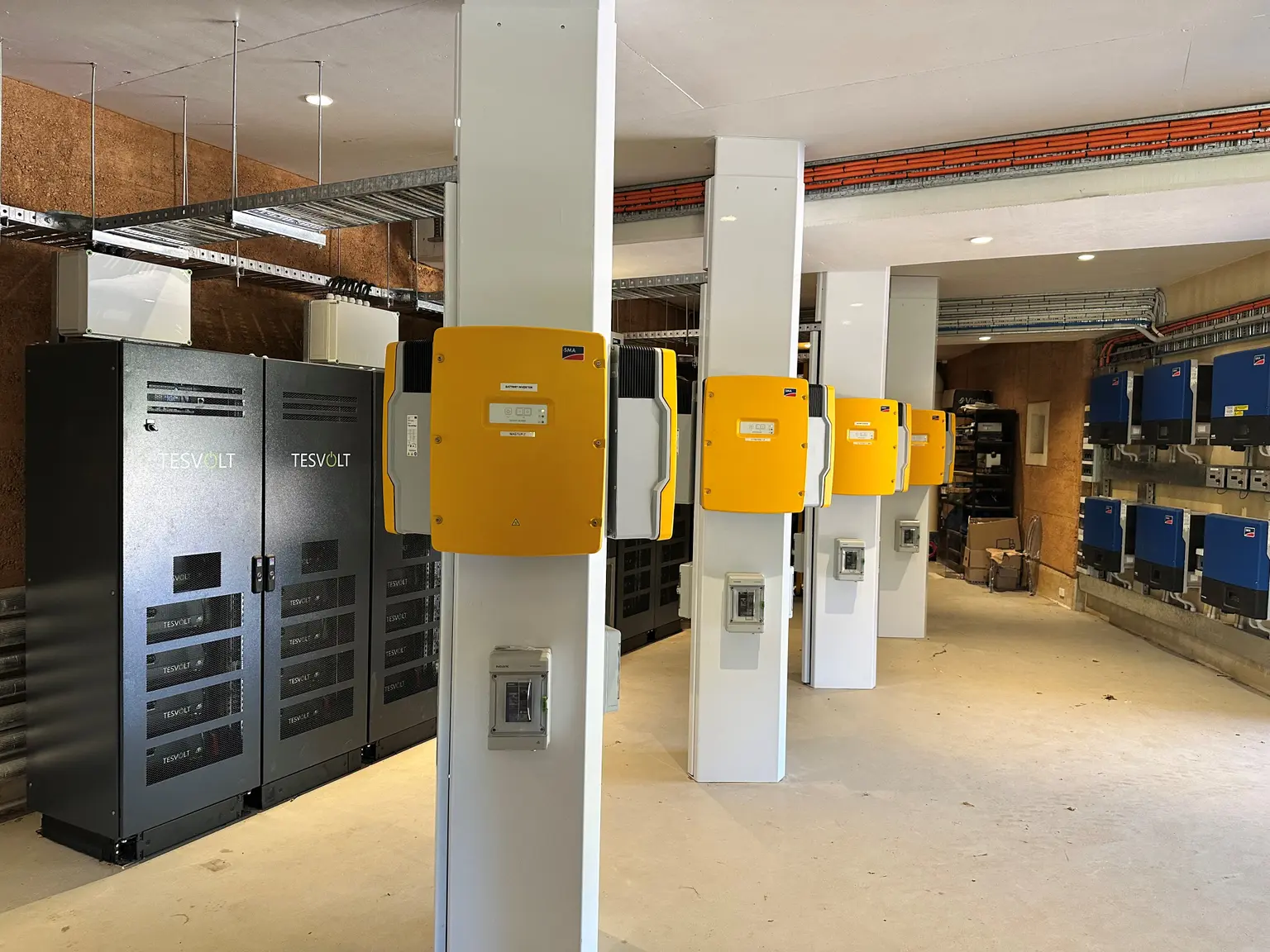 Room with industrial battery storage units and inverters on white pillars, featuring yellow and black equipment.