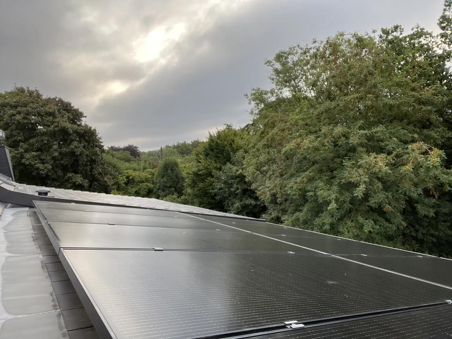 Solar panels on a rooftop under a cloudy sky, surrounded by lush green trees.