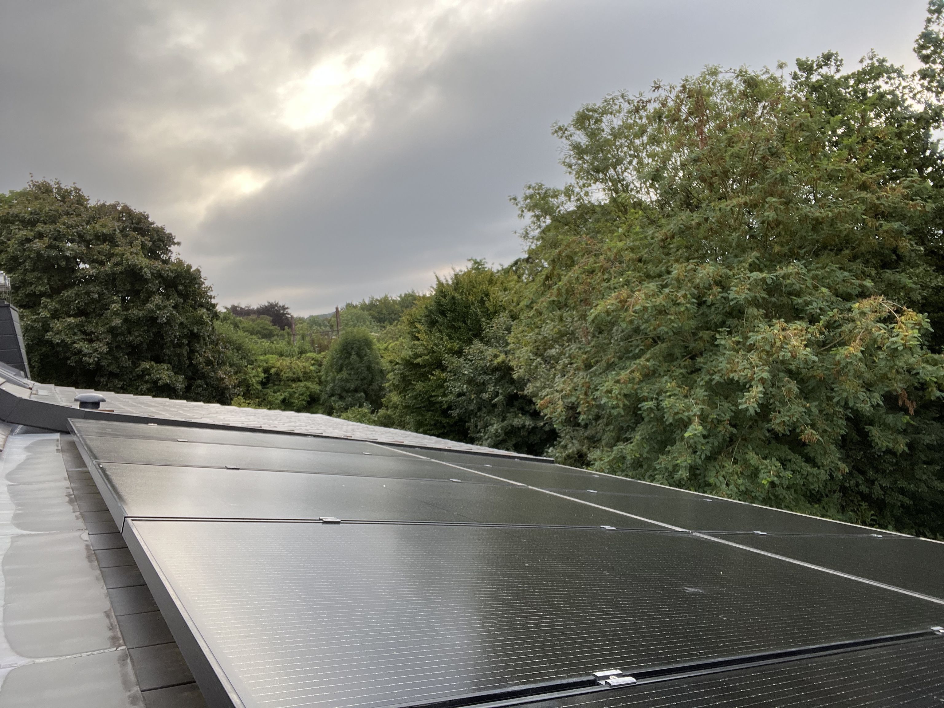 Solar panels on a rooftop under a cloudy sky, surrounded by lush green trees.