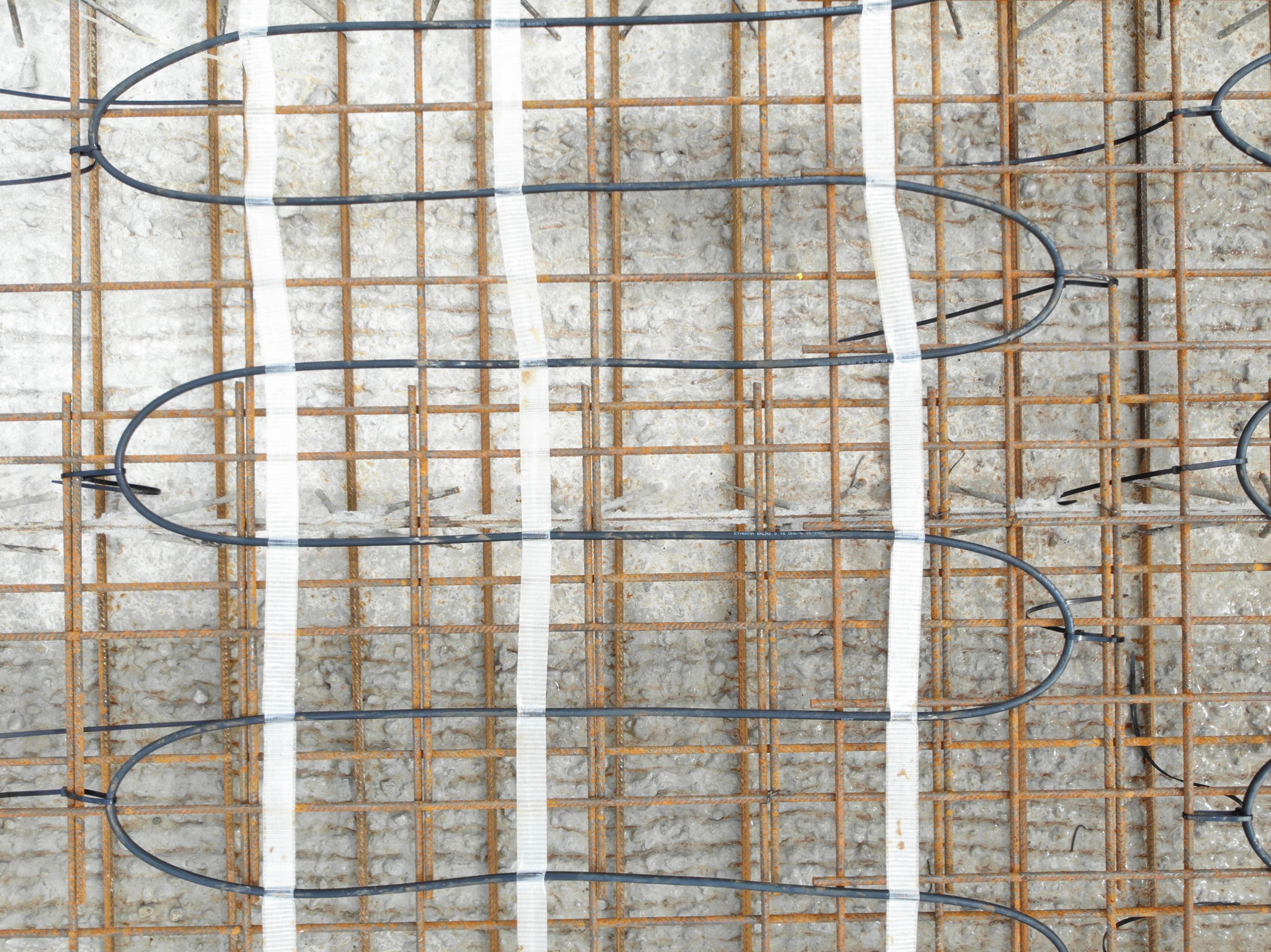 Close-up of a concrete surface with intersecting metal rebar and black tubing, likely part of underfloor heating or structural reinforcement.