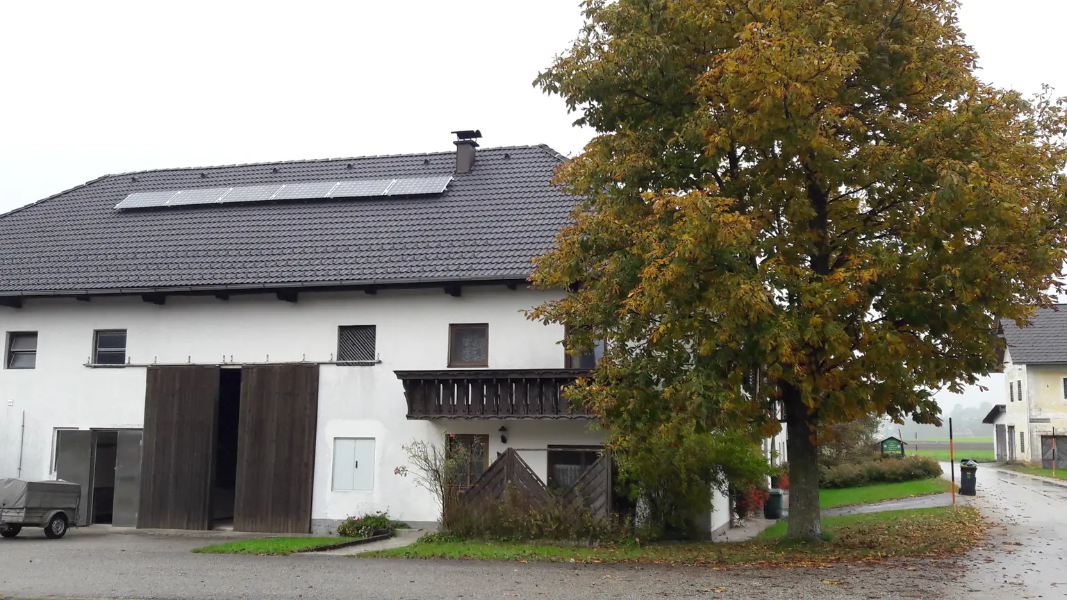 Courtyard with solar panels on dark roof, wooden balcony, and tall tree with autumn leaves in front yard.
