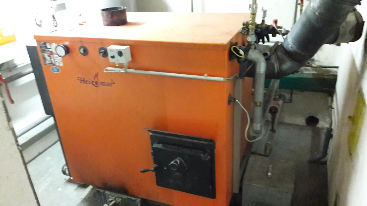 Orange industrial boiler with pipes and gauges, installed in a utility room with white walls.