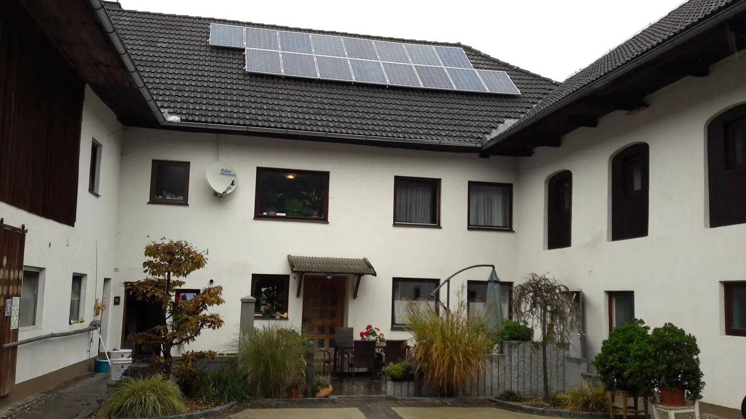 Courtyard of white building with solar panels on roof, surrounded by plants and small seating area near entrance.