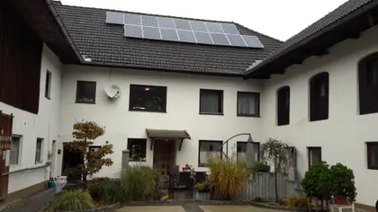 Courtyard of white building with solar panels on roof, surrounded by plants and small seating area near entrance.