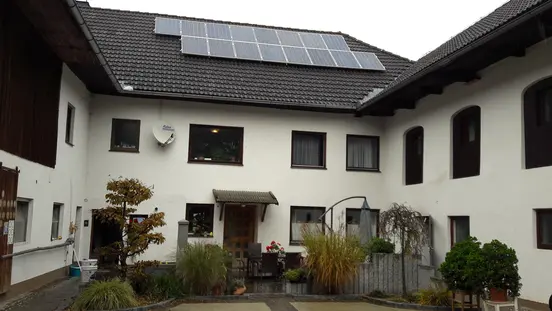 Courtyard of white building with solar panels on roof, surrounded by plants and small seating area near entrance.
