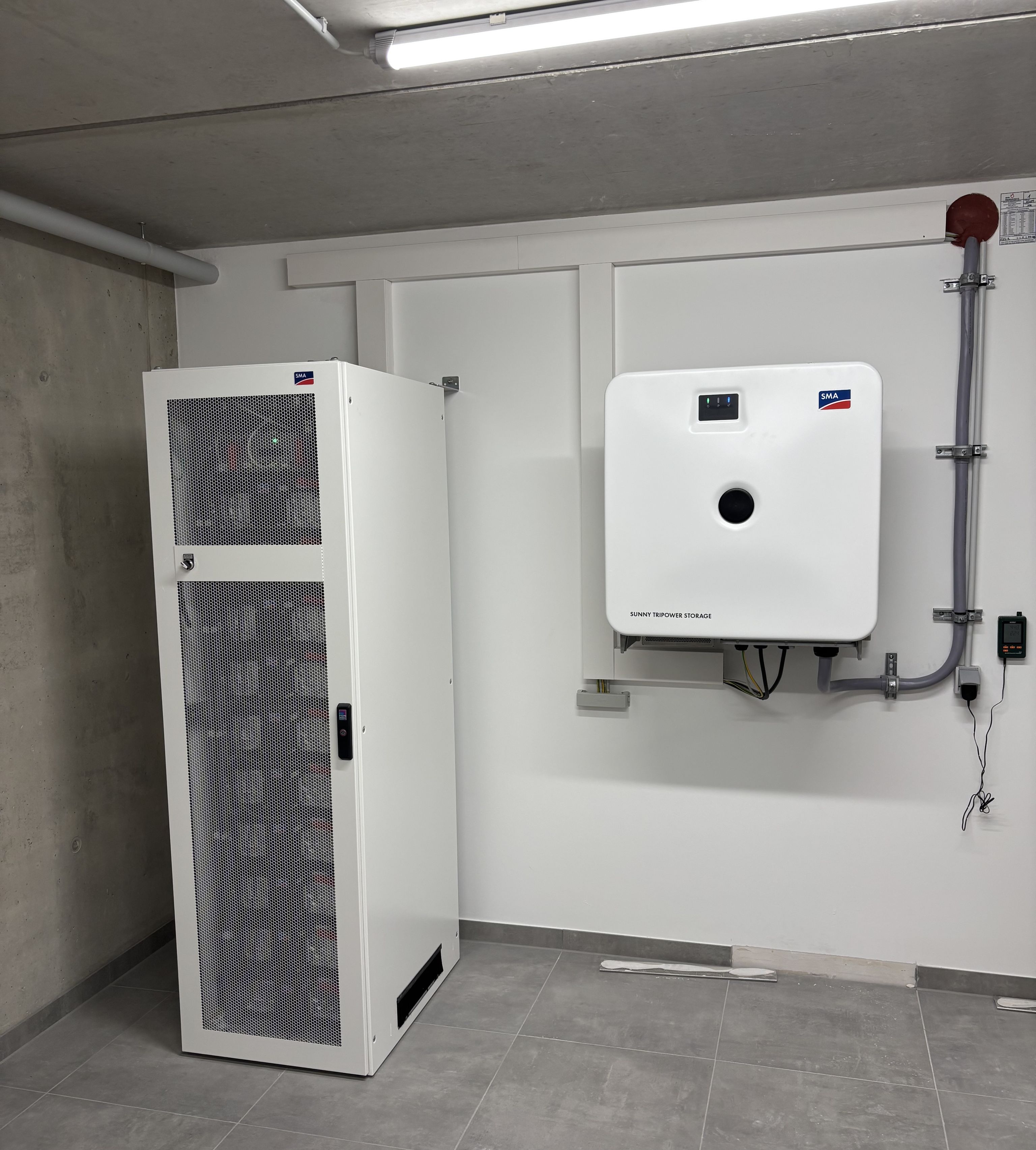 A modern electrical storage room with a tall white cabinet and a wall-mounted battery system, featuring visible pipes and a bright overhead light.