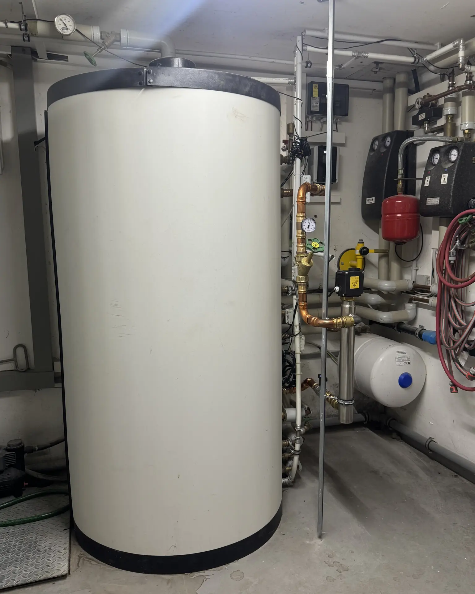 A large cylindrical water heater in a utility room, surrounded by pipes, valves, a red expansion tank, and coiled hoses against white walls.