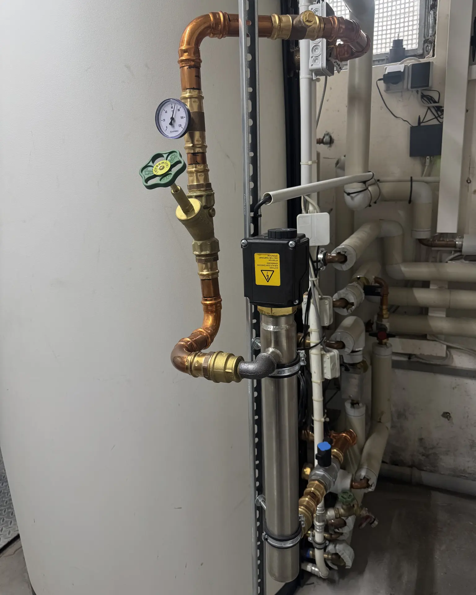 Industrial setup with pipes, valves, and a pressure gauge in a utility room. Near a large cylindrical tank.