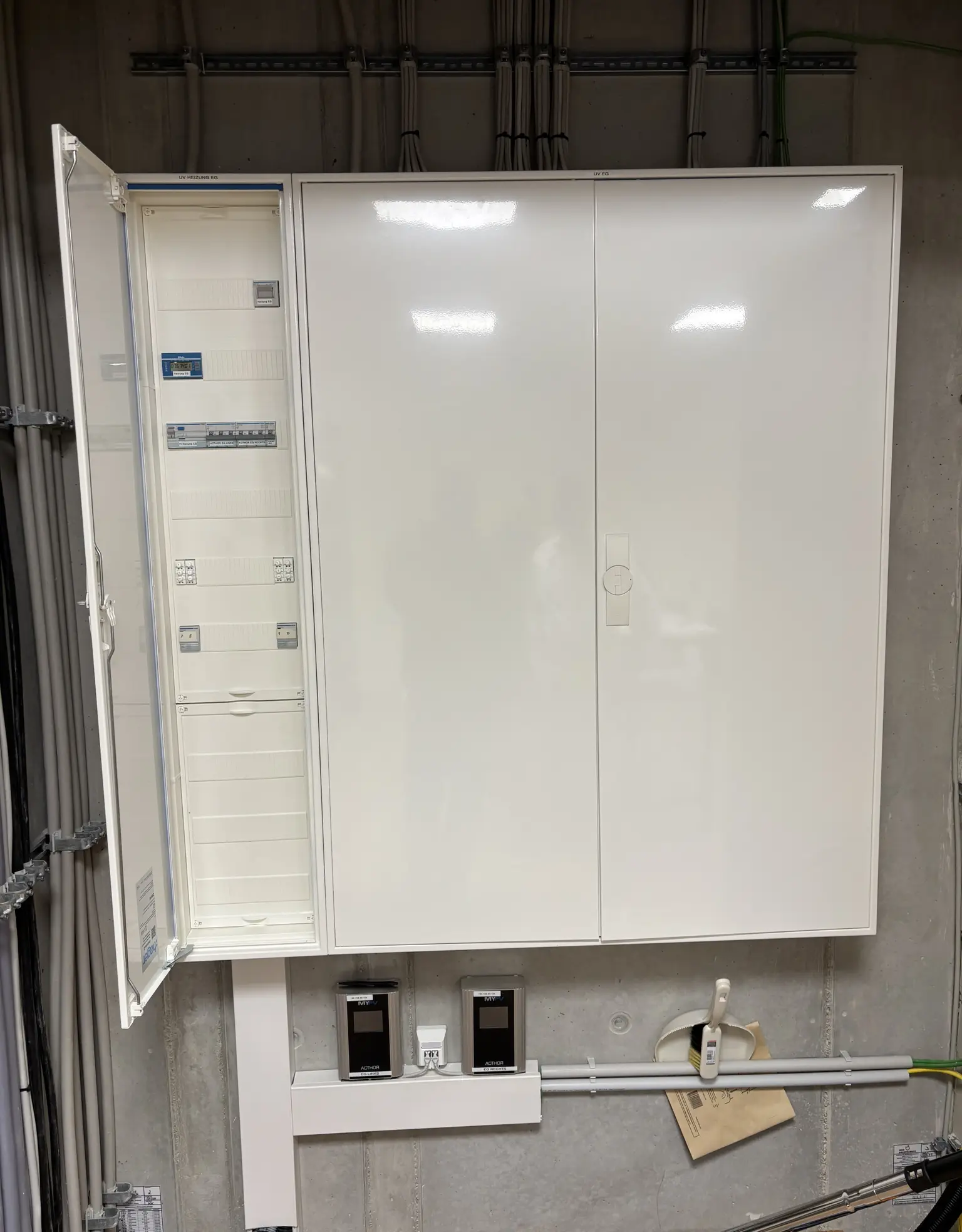 Open electrical panel with organized cabling on a concrete wall, showing circuit breakers and power meters below.
