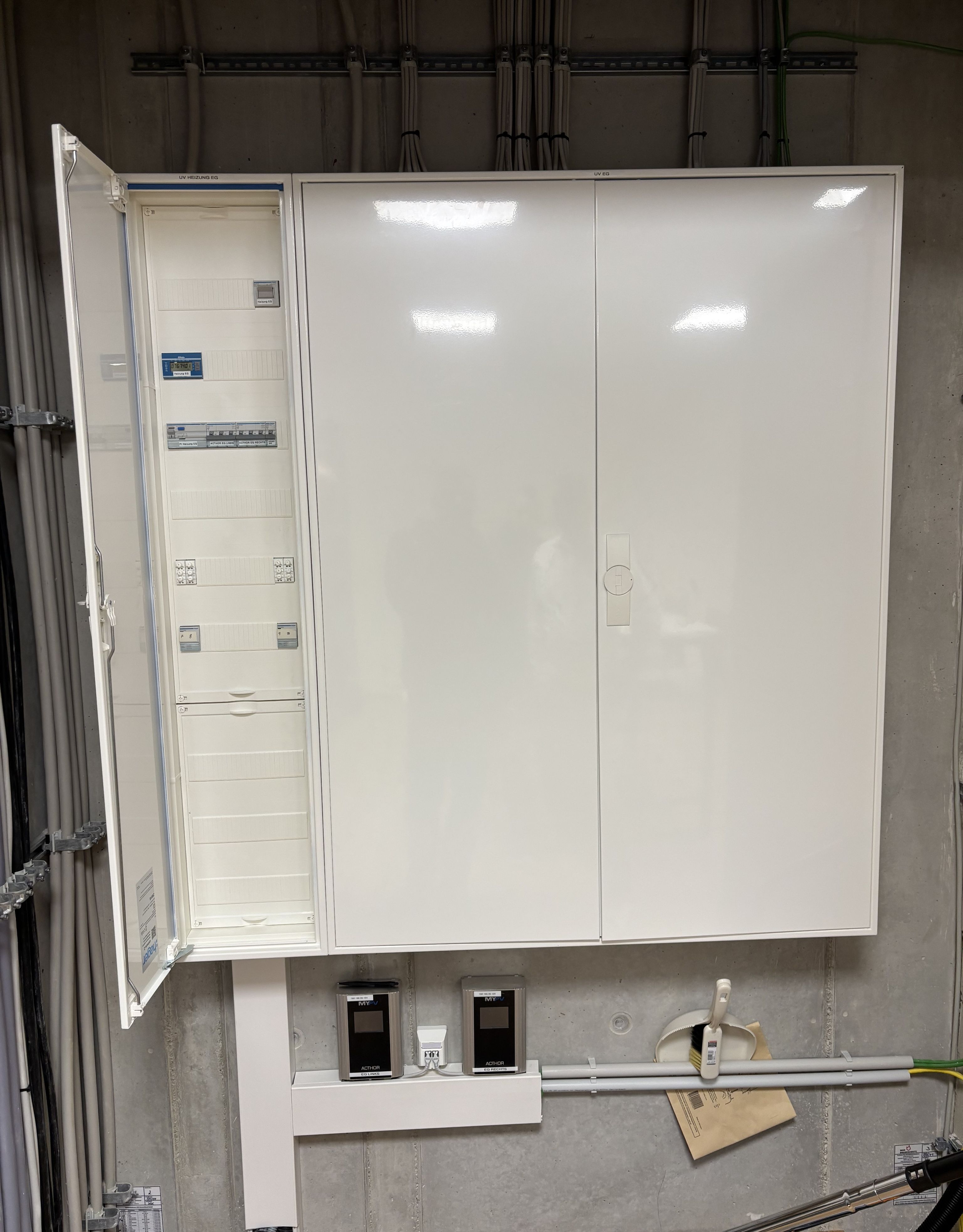 Open electrical panel with organized cabling on a concrete wall, showing circuit breakers and power meters below.