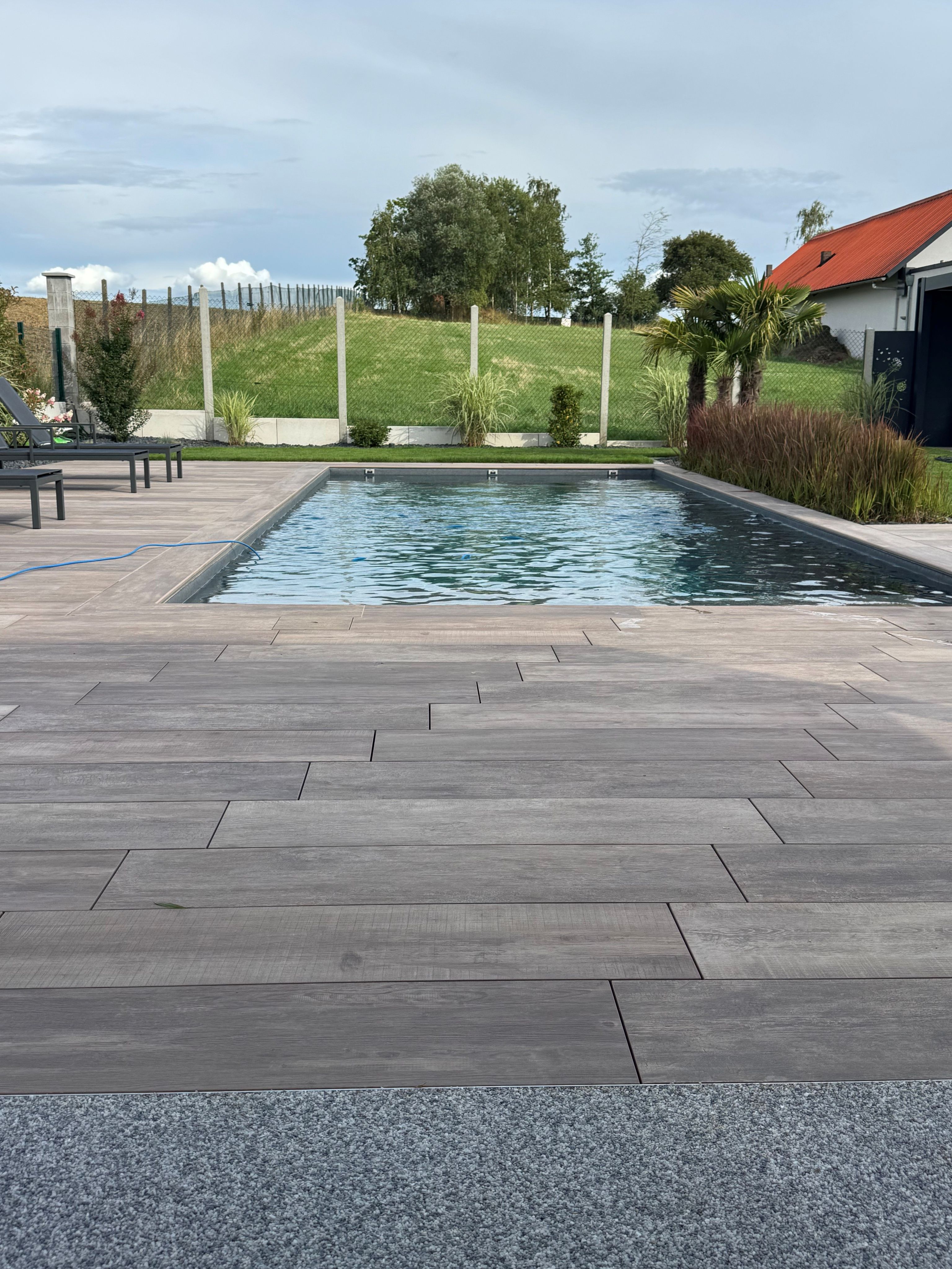 Modern backyard pool with wooden deck, surrounded by greenery, with a red-roofed house and cloudy sky in the background.