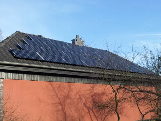 Solar panels on a sloped red house roof, with clear blue sky and bare tree branches in the foreground.