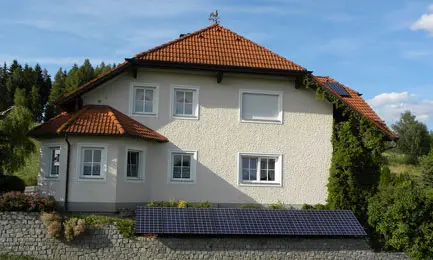 House with red-tiled roof, solar panels on roof and ground, surrounded by greenery and stone wall under blue sky.