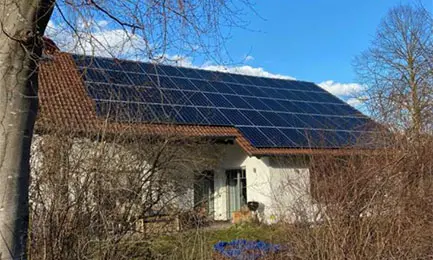 House with solar panels on the roof, surrounded by bare trees and bushes under a clear blue sky.