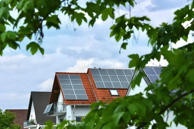 Red-tiled houses with rooftop solar panels, framed by green leaves against a cloudy sky.
