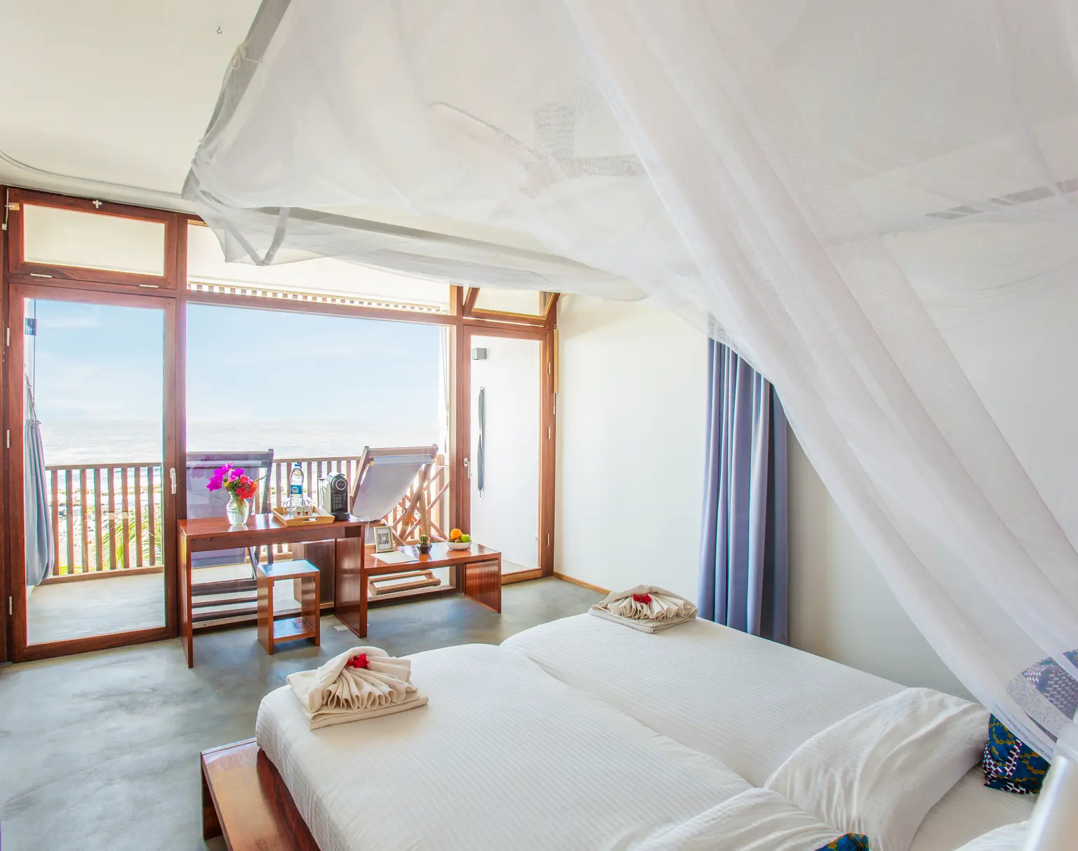Hotel room with central bed, white linens, blue pillows, towel art, and a balcony with two loungers