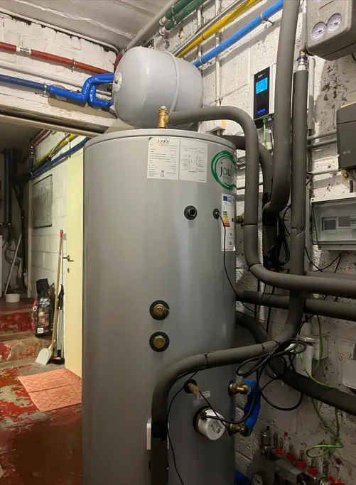 A large gray water heater with pipes and gauges is installed in a utility room with exposed brick walls and various colored pipes.