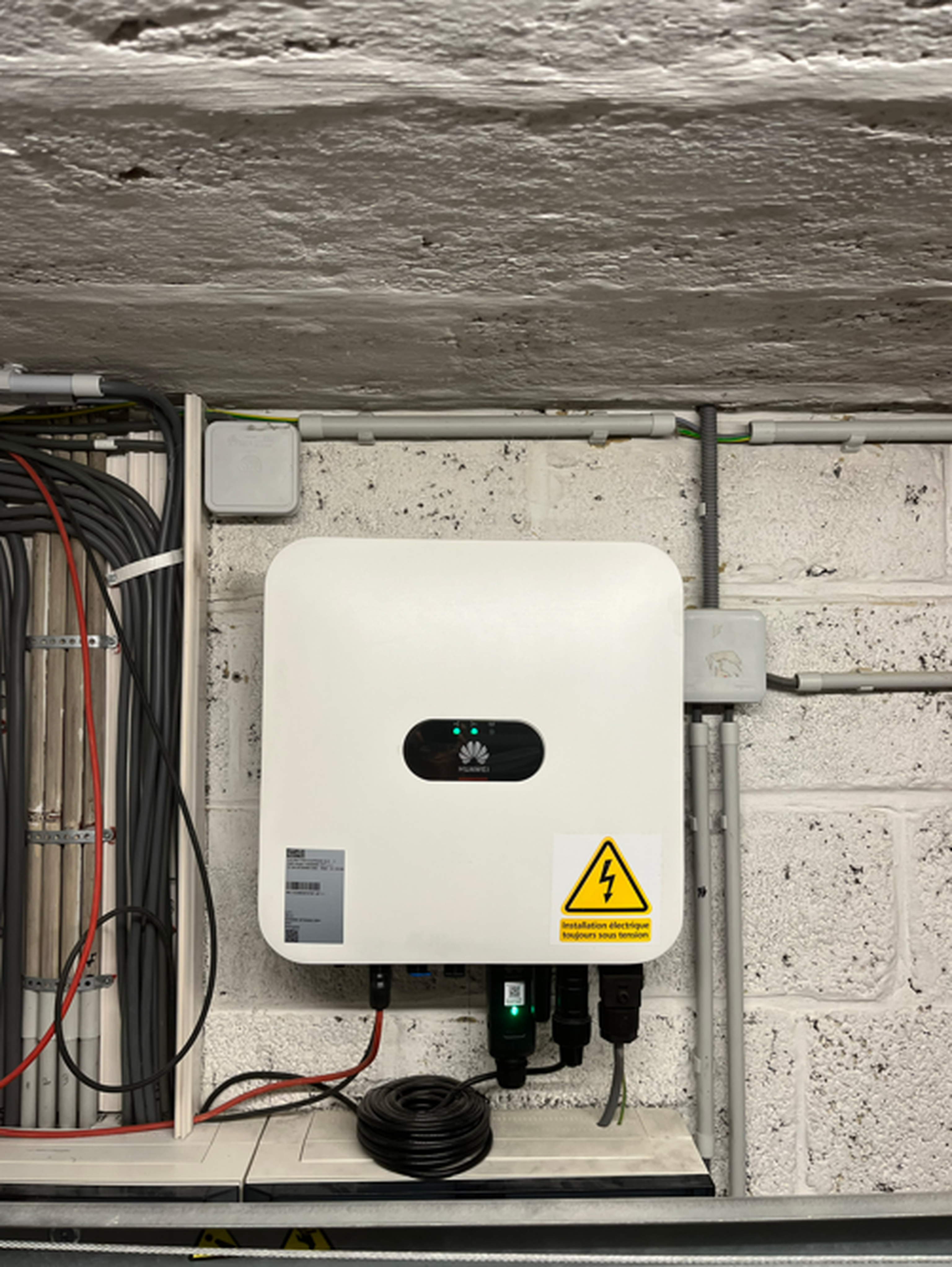 White electrical device mounted on a brick wall, surrounded by cables, with a caution sign indicating high voltage.
