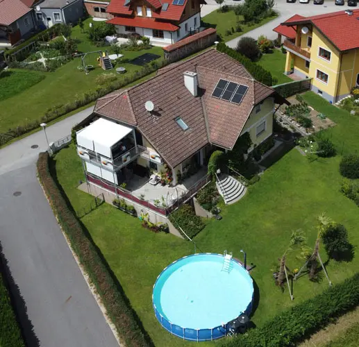 Aerial view of a suburban house with a brown roof, solar panels, garden, and a circular above-ground pool in the backyard.