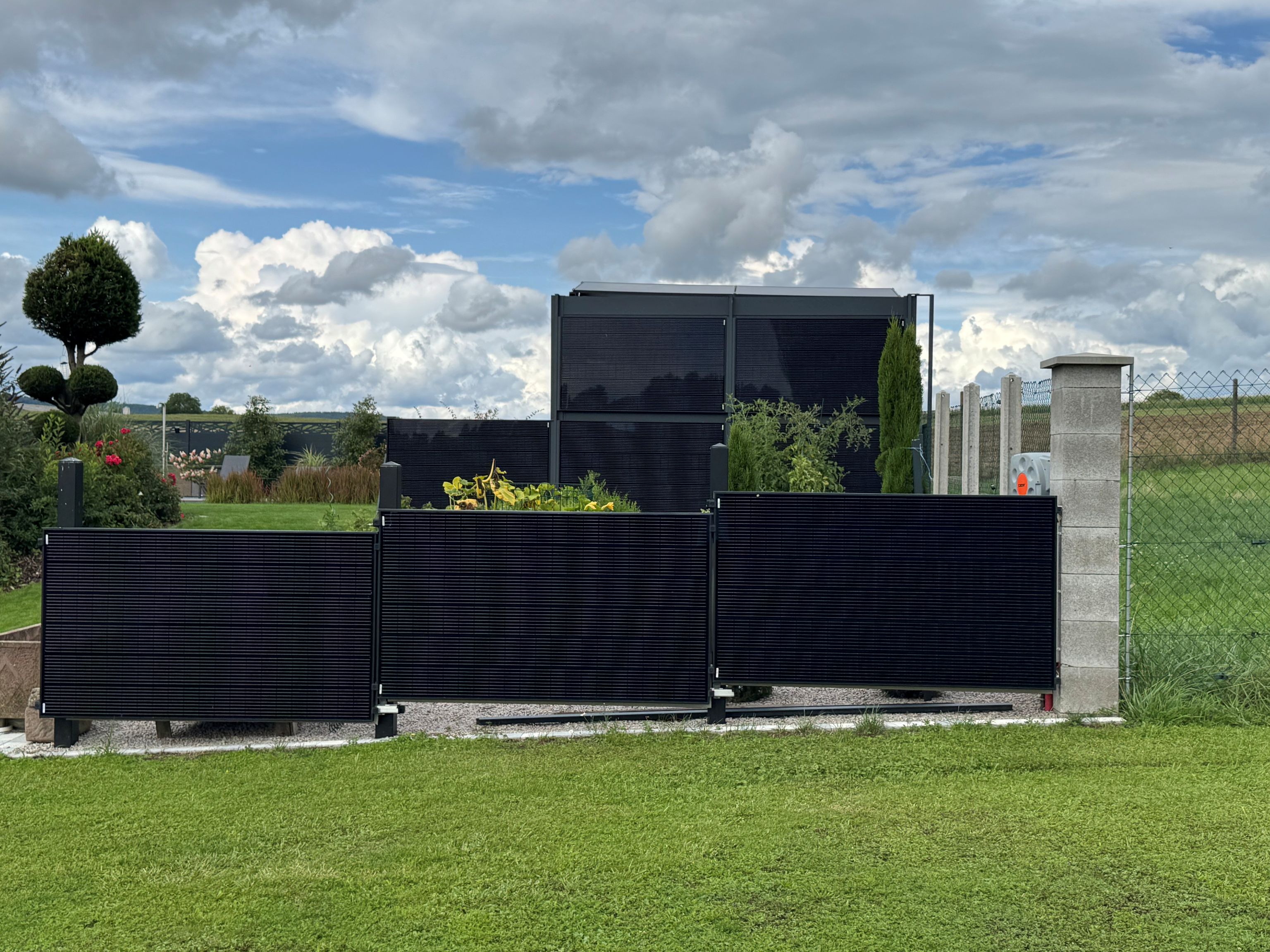 Modern garden with solar panel fence, lush greenery, and a cloudy sky in the background.
