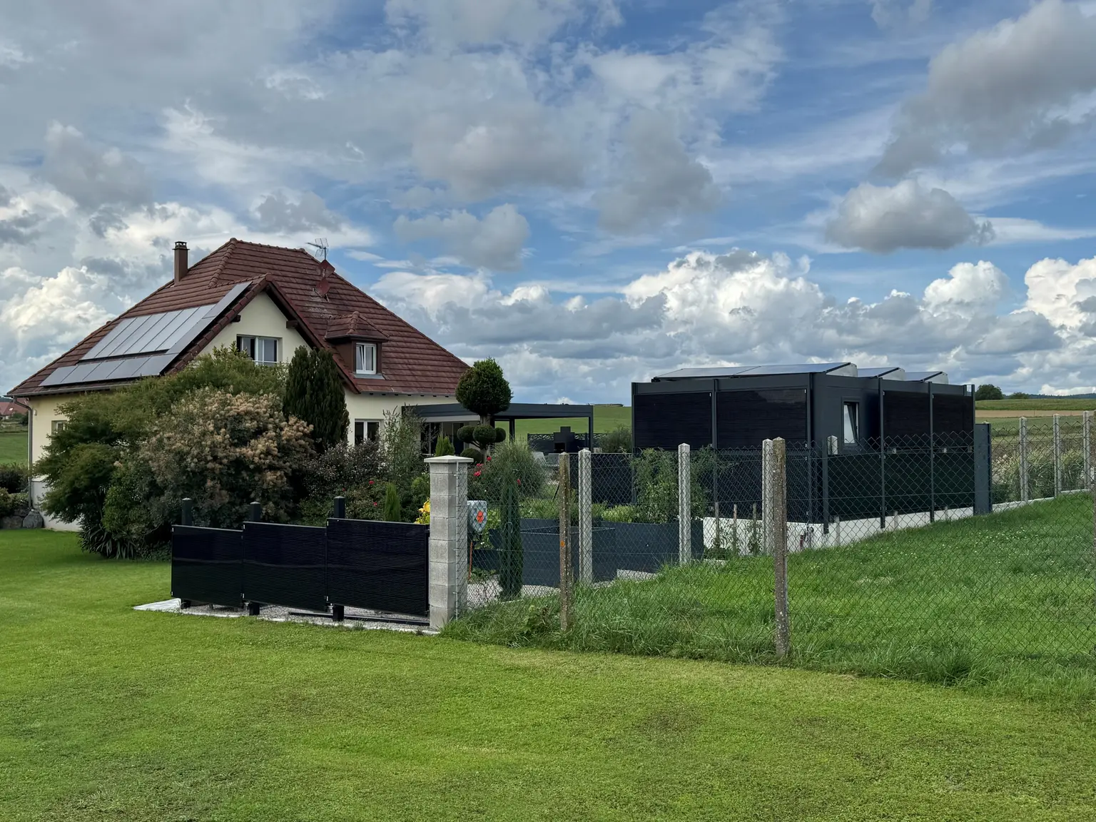 House with solar panels and garden next to fenced area, surrounded by green grass under a cloudy sky.