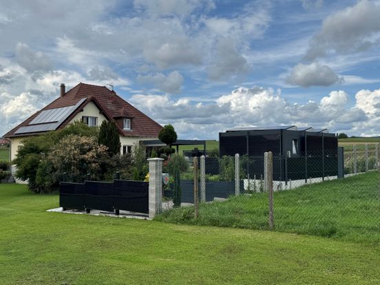 House with solar panels and garden next to fenced area, surrounded by green grass under a cloudy sky.