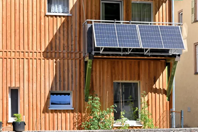 Wood-paneled building facade with a balcony-mounted array of solar panels, two windows and potted plants at the base.