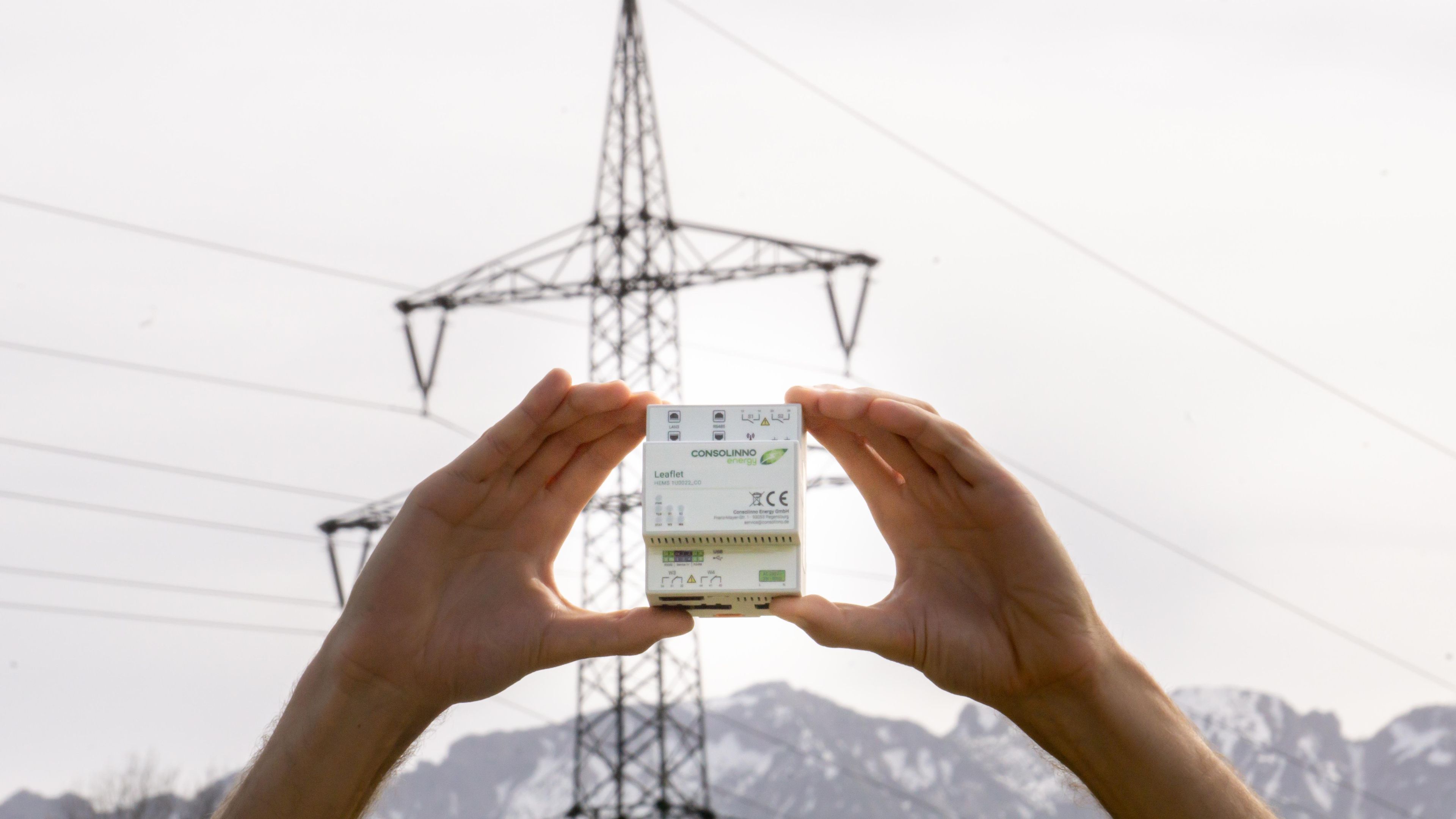 Hands hold a small electronic device against a backdrop of power lines, with mountains and a clear sky in the background.