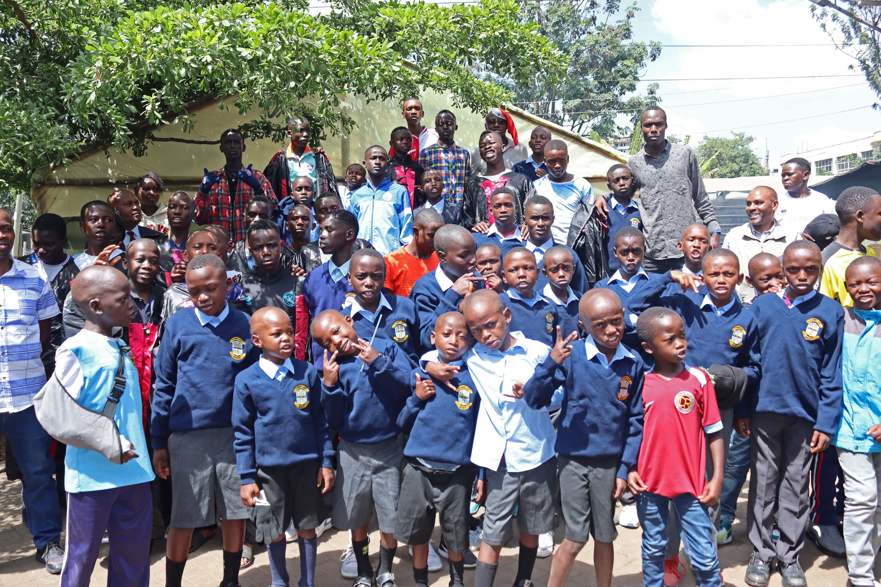 A large group of children in school uniforms and adults gather outdoors, smiling and posing for the photo. Trees and a building are in the background.