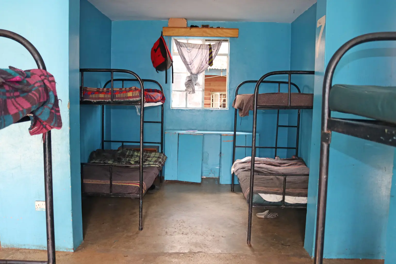 Simple room with blue walls, containing three bunk beds, a window with cloth drapes, and a small shelf holding various items.