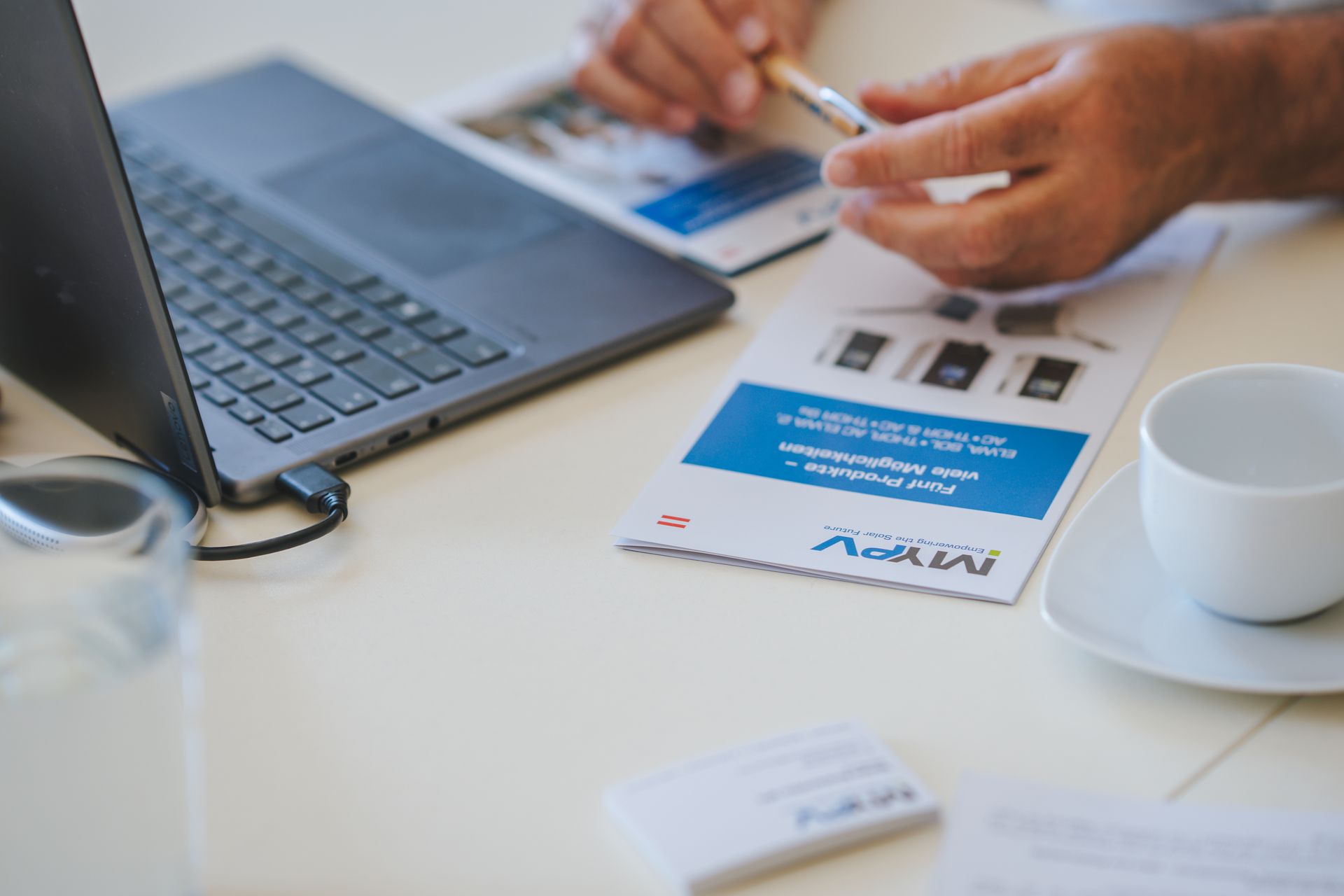 A person holding a pen reviews a brochure next to a laptop and a white coffee cup on a desk.