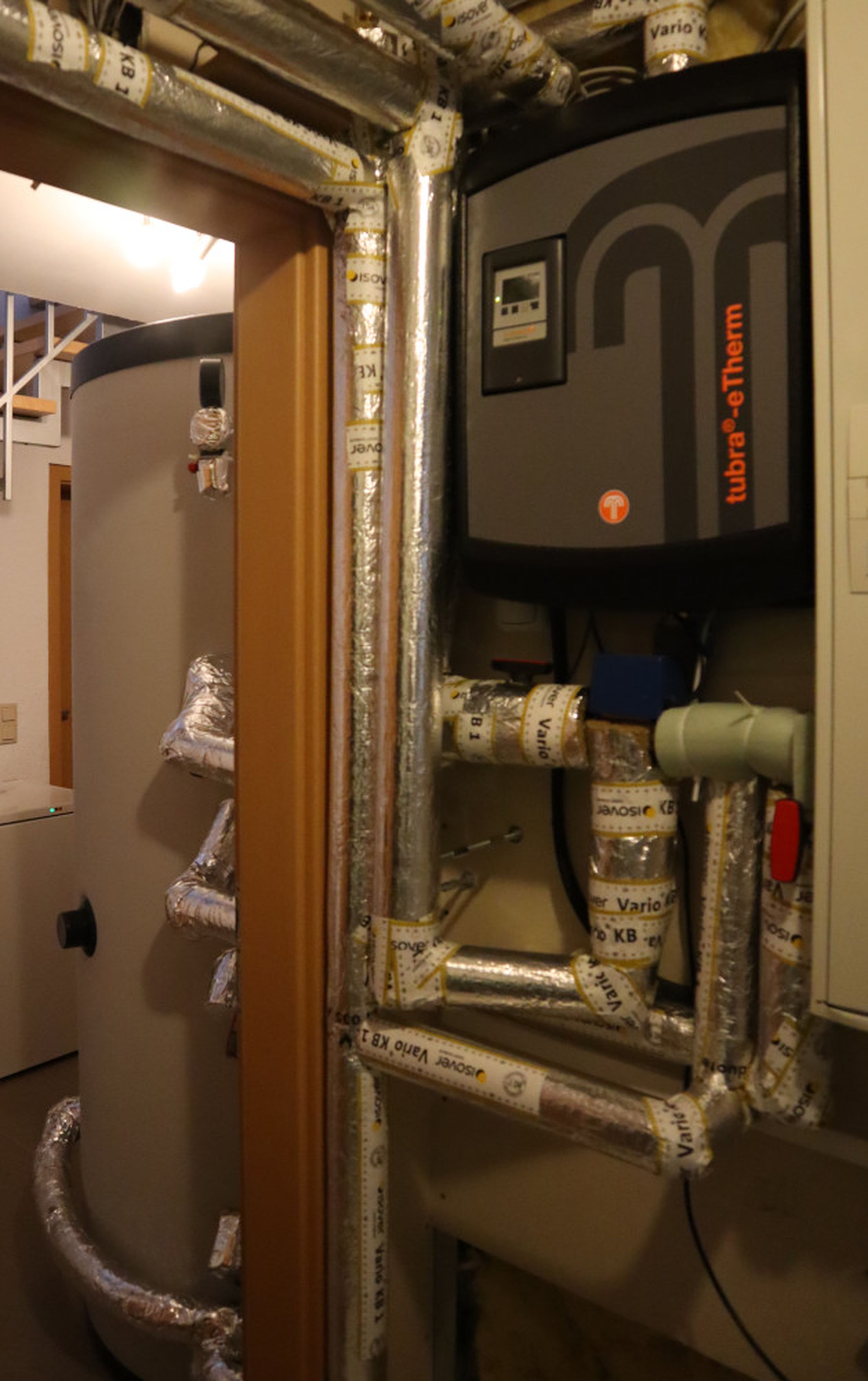Basement boiler room with a large water heater, insulated pipes, and a control panel mounted on the wall, connected by labeled ducts.