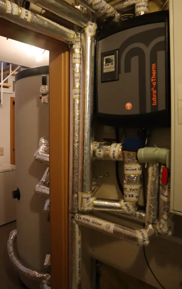 Industrial boiler room with insulated piping, a large cylindrical heater tank, and a control panel mounted on the wall.
